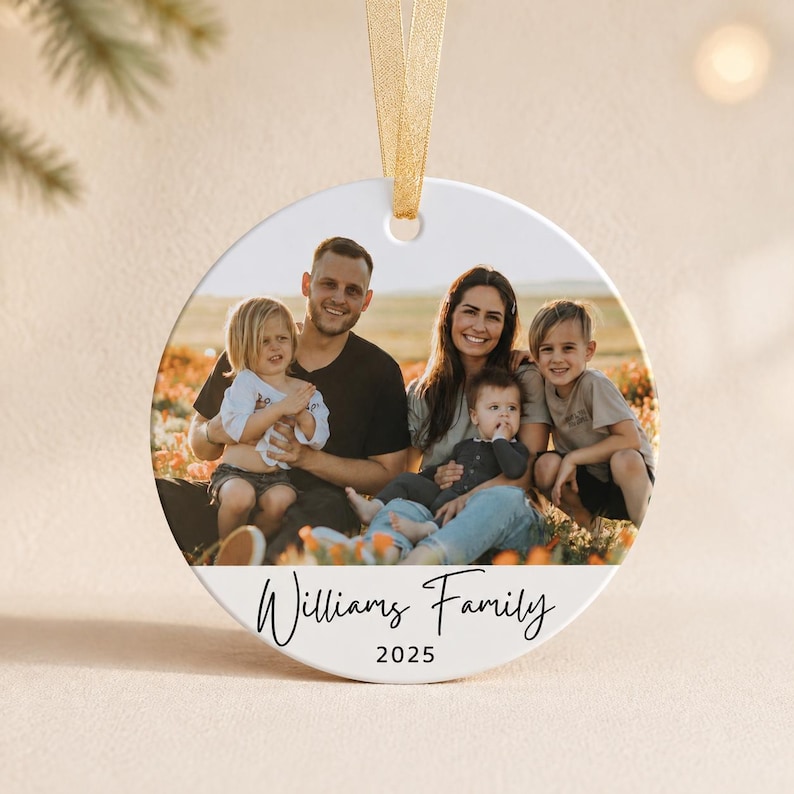Puede incluir: Un adorno redondo de cer&aacute;mica blanca con una fotograf&iacute;a de una familia de cuatro personas sentadas en un campo. El texto "Williams Family" est&aacute; impreso en negro bajo la foto, con el a&ntilde;o "2025" debajo. Una cinta dorada est&aacute; sujeta en la parte superior.