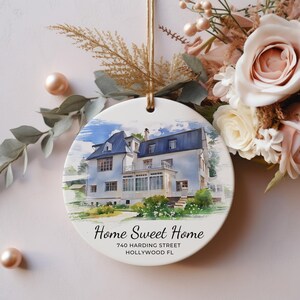 Puede incluir: Un adorno redondo de cer&aacute;mica blanca con una ilustraci&oacute;n en acuarela de una casa de dos pisos con techo oscuro. El texto dice Home Sweet Home, 740 Harding Street, Hollywood FL. Cuelga de una cinta dorada y est&aacute; rodeado de detalles florales.