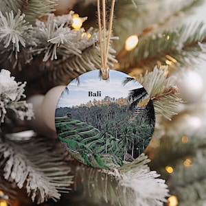 May include: A round ceramic ornament with a picture of rice paddies in Bali, Indonesia. The ornament has a brown twine hanger and the word "Bali" printed in black on the front.