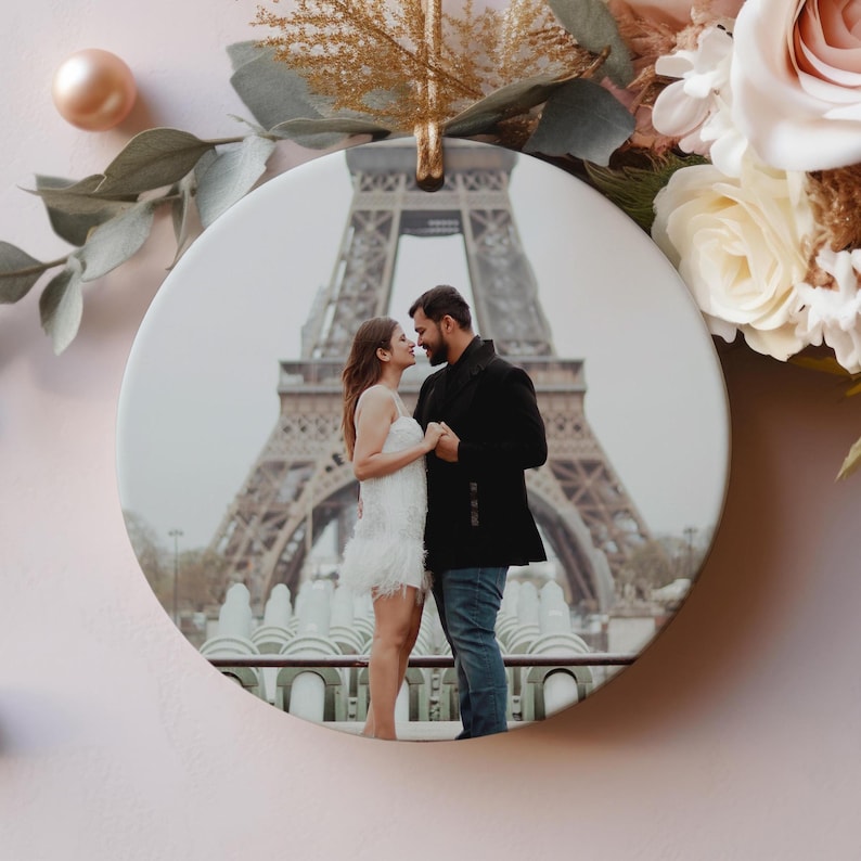 Puede incluir: Un adorno redondo de cer&aacute;mica con una fotograf&iacute;a impresa de dos personas frente a la Torre Eiffel. Una persona viste un vestido blanco y la otra una chaqueta negra y pantalones vaqueros azules. El adorno est&aacute; decorado con flores artificiales.