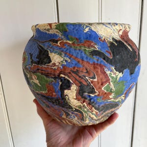 May include: A round ceramic bowl with a marbled design. The bowl features a mix of colors, including blue, red, black, green, and cream. The bowl is held in a hand, showcasing its size and shape.