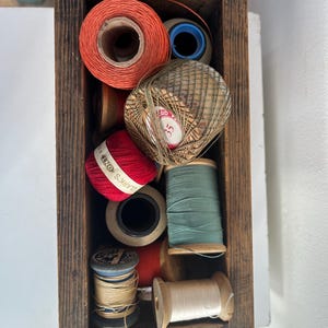 May include: A wooden box filled with spools of thread in various colors, including red, white, green, and beige. The spools are arranged in a random pattern and some have labels with text.