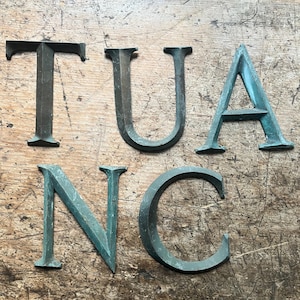 May include: Five individual metal letters forming the sequence T, U, A, N, and C. Each letter features a weathered, oxidized patina with shades of dark green and bronze. The letters are constructed with a serif typeface and have a dimensional profile.