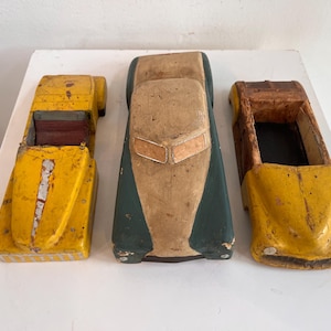 May include: Three vintage toy cars made of wood. The left car is yellow with a red interior. The center car is dark green and beige with a closed roof. The right car is yellow and brown with an open top. All three show signs of wear and age.