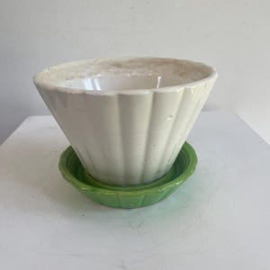 May include: A white, fluted ceramic planter with a wide, cone-like shape, sitting on a small, round, green saucer. The planter has a slightly aged appearance around the rim. The set is designed for indoor or outdoor use.