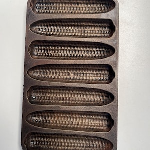 May include: A cast iron corn on the cob pan with six individual molds. The pan is dark brown and has a rustic look.