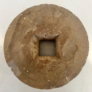 May include: A circular, weathered stone object with a rough, textured surface in shades of tan and brown. The object features a prominent, centered square-shaped hole that extends through the middle, creating a distinct geometric opening.