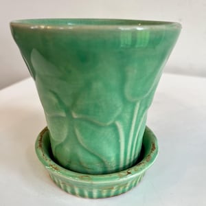 May include: A green ceramic planter with a matching saucer. The planter has a flared shape and a textured leaf design. The saucer has a ribbed edge. The planter and saucer are a light green color.