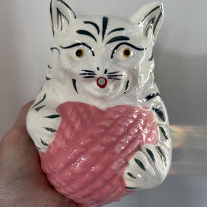 May include: White ceramic cat figurine with black stripes and green accents. The cat is holding a pink ball of yarn in its paws.