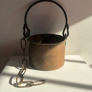 May include: A copper bucket with a metal handle and chain. The bucket has a worn, aged look and is perfect for rustic decor.