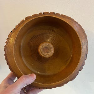 May include: A handcrafted wooden bowl with a natural edge. The bowl is a warm brown color, with a circular design in the center. The bowl is held by a hand.
