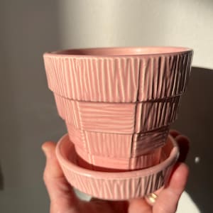 May include: A pink ceramic planter with a matching saucer. The planter has a tiered design with vertical and horizontal textured lines. The saucer is round with similar textured lines. The planter is held in a hand.