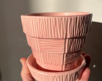May include: A pink ceramic plant pot with a matching saucer. The pot has a tiered design with vertical and horizontal textured lines. The saucer is round with similar textured lines. The pot is held in a hand.