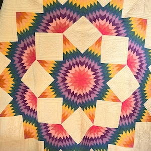 May include: A quilt featuring a large central star pattern composed of small diamond-shaped fabric pieces in a gradient of pink, purple, blue, yellow, and orange. The star is set against a cream-colored background with intricate quilted stitching.