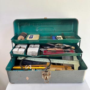 May include: A vintage metal cantilever tool box with a grey base and a green interior. The tiered trays contain various items including white blocks, a spool of wire, a metal ruler, pencils, a paintbrush, and a small red-lined case.