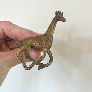 May include: A small, metallic, brass-colored giraffe figurine with a textured surface. The giraffe is depicted in a running or leaping pose with its legs bent and its head turned slightly to the right, showing a weathered, aged patina finish.
