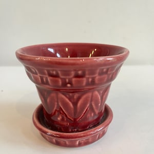 May include: A burgundy ceramic flower pot with a matching saucer. The pot has a decorative embossed pattern and a wide rim. The saucer is round and fits snugly beneath the pot. The pot is designed for small plants.