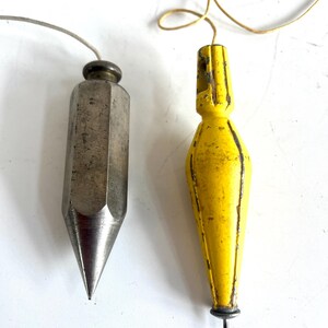 May include: Two plumb bobs, one silver-colored and the other yellow. The silver plumb bob is a faceted, elongated cone shape with a pointed tip and a screw top. The yellow plumb bob is a teardrop shape with a loop at the top and a small metal pin at the bottom.