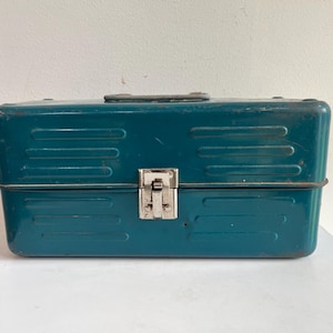May include: A rectangular teal metal storage box featuring a horizontal ribbed design on the front and back panels. The container includes a central silver-colored metal latch mechanism and a dark handle attached to the top lid surface.