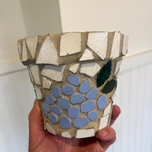 May include: A mosaic-style flower pot with a light blue grape design and green leaves. The pot is made of white and light gray tiles. The pot is designed to hold plants.