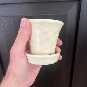 May include: A small, off-white ceramic pot with a drainage hole and a matching saucer. The pot has a textured surface.