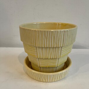 May include: A yellow ceramic planter with a matching saucer. The planter has a textured, woven basket design. The color is a soft, pale yellow. The planter is designed for indoor or outdoor use.