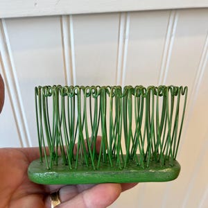 May include: A green wire flower frog featuring a rectangular base with numerous vertical wire loops arranged in a dense, upright pattern. The metal structure is painted in a solid green finish, designed to hold stems in place for floral arrangements.