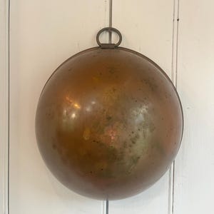 May include: A round, polished copper bowl with a smooth, reflective surface and a weathered patina. The bowl features a small, circular metal ring attached to the top rim, designed for hanging. The copper has subtle color variations and dark spots.