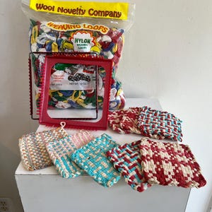 May include: A collection of weaving supplies, including a bag of nylon weaving loops, a red weaving loom, and several woven pot holders in various colors. The bag reads "Wool Novelty Company" and "Weaving Loops".