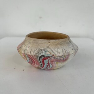 May include: A small, white ceramic bowl with a marbled design in shades of pink, red, blue, and black. The bowl has a rounded bottom and a slightly flared rim.