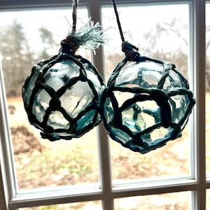 May include: Two clear glass fishing floats encased in teal netting, suspended by twine. The floats are round and have a slightly aged appearance. The background shows a window with a view of trees.