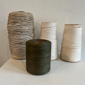 May include: Four spools of twine in varying colors and sizes. The largest spool is a light tan color, while the others are white and olive green. The spools are arranged on a white surface against a neutral background.
