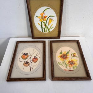 May include: Set of three framed watercolor paintings. Each painting features a different floral or botanical subject within an oval frame. The frames are dark brown wood. The paintings include orange flowers, persimmons, and yellow flowers.