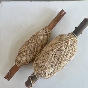 May include: Two handmade yarn winders. Each winder features a wooden handle and is wrapped with natural-colored twine. The twine is wound around the wooden handle in a symmetrical pattern. The winders are displayed on a white surface.