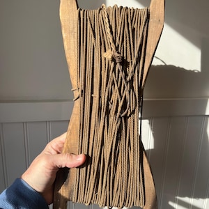 May include: A vintage wooden rope winder with a spool of brown rope. The winder has a rectangular frame with two upright handles and two feet. The rope is tightly wound around the frame.