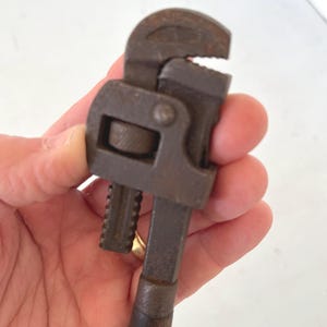 May include: A vintage pipe wrench with a dark brown wooden handle and a rusty, metal head. The adjustable jaws are open, revealing the teeth. This tool is designed for gripping and turning pipes.