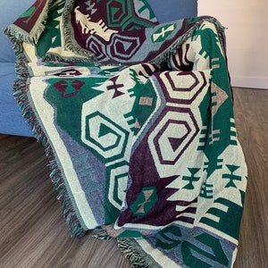 Peut inclure: Une couverture en laine tissée de couleur verte, blanche et violette avec un motif géométrique. La couverture a des franges sur tous les côtés et est drapé sur un canapé bleu.
