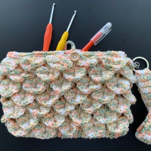 Peut inclure: Un sac en crochet avec un motif d'écailles en blanc, vert et pêche. Le sac a une poignée et une fermeture à bouton. Trois crochets à crochet et un stylo sont à l'intérieur du sac.
