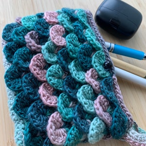 Peut inclure: Une pochette en crochet avec un motif texturé en forme d'écailles dans des tons de bleu sarcelle, vert et rose. La pochette a un bouton noir et une boucle pour une sangle.