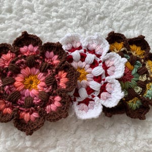 May include: Three handmade crochet flower coasters in shades of pink, red, white, yellow, and brown. The coasters are arranged on a white, textured surface. Each coaster has a unique color palette and floral design.