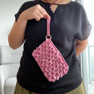 Peut inclure: Un sac à main en crochet rose avec une bandoulière en chaîne. Le sac a un motif en forme d'écailles.