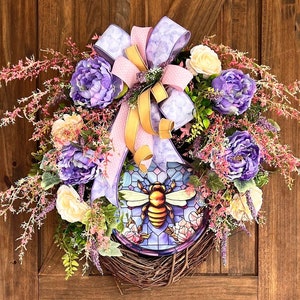 Può includere: Una ghirlanda floreale viola e rosa con un designo di ape in vetro colorato. La ghirlanda è realizzata con fiori e verde artificiali e ha una base in vimini marrone.