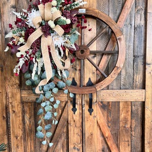 Può includere: Una ruota di carro in legno decorativa ornata da una composizione floreale con rose rosso scuro, dalie bianche, foglie di eucalipto verdi e rametti di felce bianchi, arricchita da un grande fiocco in juta color sabbia.