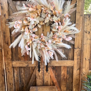 Può includere: Una grande ghirlanda circolare con un mix di erba della pampa finta, foglie di eucalipto e delicati accenti floreali rosa e beige. La ghirlanda è esposta contro una porta di legno rustica, creando un'estetica calda e invitante. Decorazione.