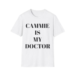 May include: White t-shirt with the text "CAMMIE IS MY DOCTOR" in black, bold letters. The shirt has a crew neck and short sleeves. The text is centered on the front of the shirt.