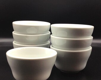May include: A set of eight white ceramic bowls, stacked in two piles. The bowls are smooth and have a slightly rounded shape.