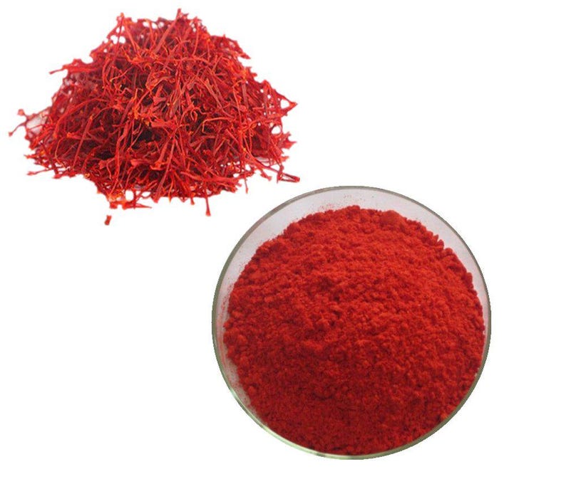 May include: A pile of vibrant red saffron threads next to a small glass bowl filled with red saffron powder. The saffron threads are long and thin, while the powder is finely ground. The background is plain white.