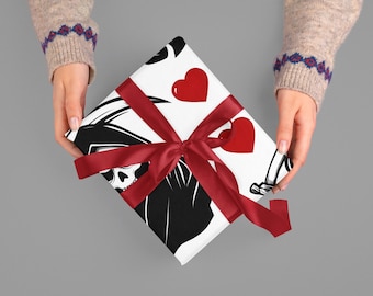 May include: A wrapped gift with a white background featuring a black Grim Reaper design and red hearts. The gift is tied with a red satin ribbon. The hands holding the gift are wearing a beige sweater.