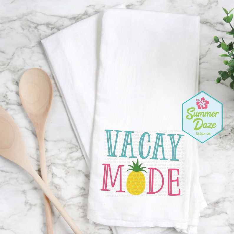 May include: White kitchen towel with the text "Vacay Mode" in blue and pink lettering with a yellow pineapple graphic. The text "Summer Daze Design Co." is in a hexagon shape with a pink hibiscus flower.