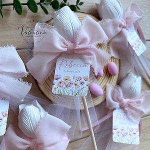 K&ouml;nnte beinhalten: Wei&szlig;e tulpenf&ouml;rmige Gastgeschenke mit rosa Schleifen und Etiketten mit der Aufschrift "Rebecca" und "4 Ottobre 2025". Die Gastgeschenke befinden sich auf Holzst&auml;ben und sind mit rosa T&uuml;ll verziert. Die Gastgeschenke befinden sich auf einer Holzscheibe.